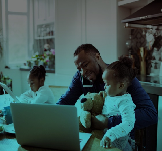 Working from home father multitasking with a child on his lap while another child sits on the side using an ipad.