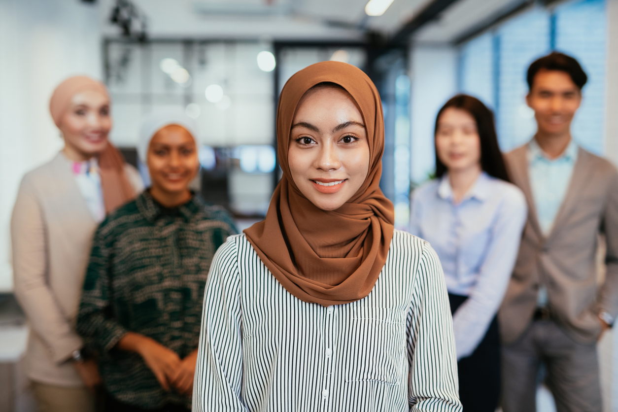 Diverse team of Asian professionals with a Muslim woman in the foreground wearing a hijab, standing in a modern office environment.