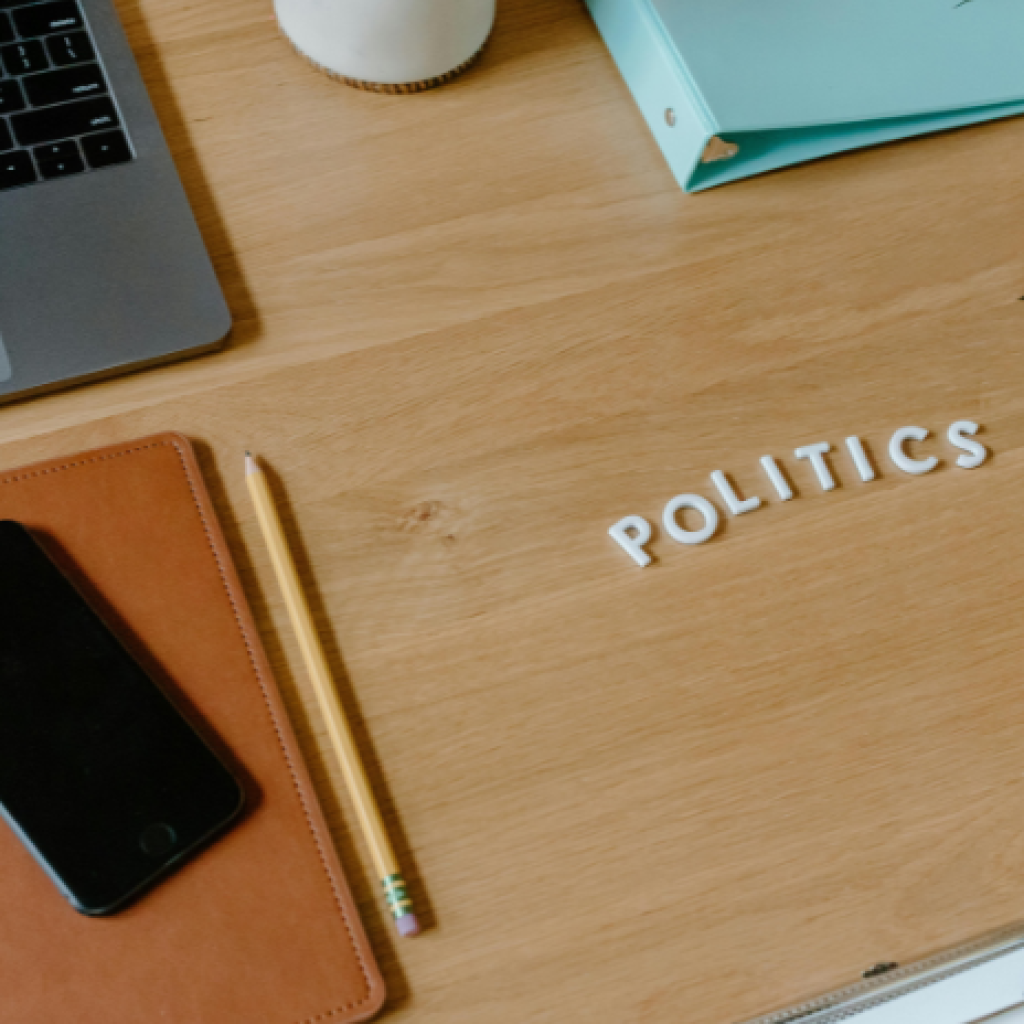 A work desk holding a laptop, phone, notebook,files and a pen - as well as small letter spelling out 'politics'