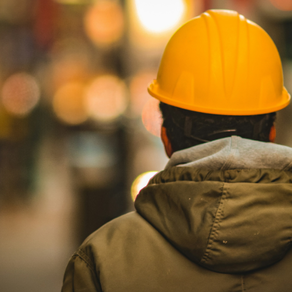 A lone worker picture from behind from the shoulders up, wearing a hooded jacket and a hardhat. The setting beyond the worker is out of focus