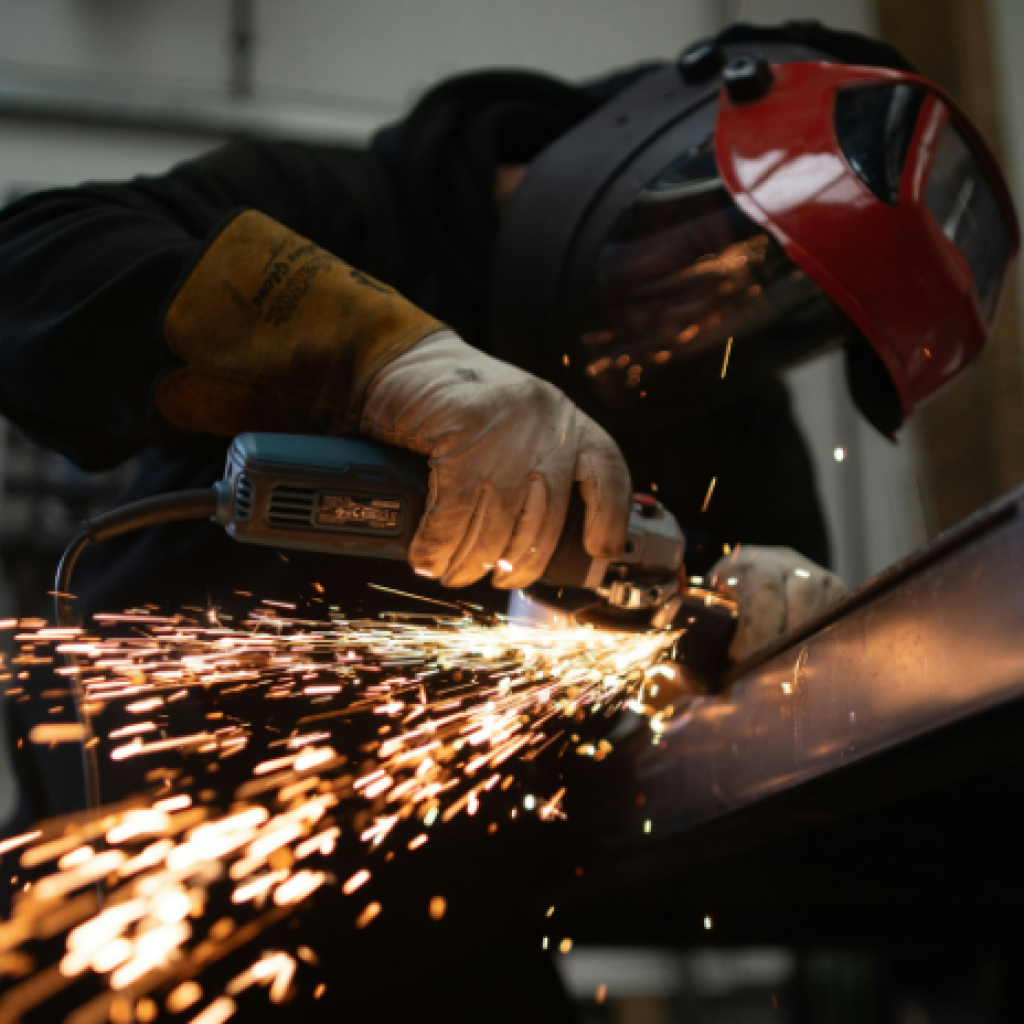 A manufacturing professional sawing through a sheet of metal, with sparks flying towards the bottom-left side of the image
