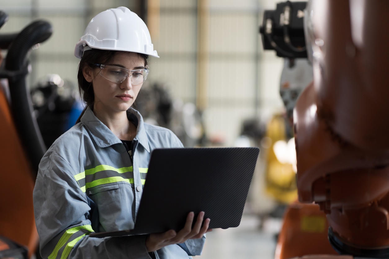 Industrial Manufacturing Robotic Arms Maintaining. Female engineer holding and working with laptop computer inspecting quality parts of robotics arms for manufacturing lines in robotic assembly plant