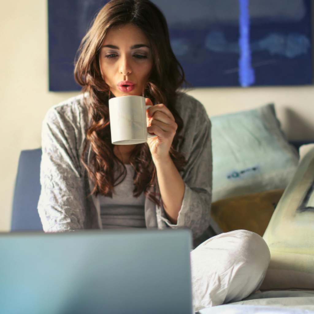 A young woman holding a cup of tea, working on her laptop in bed