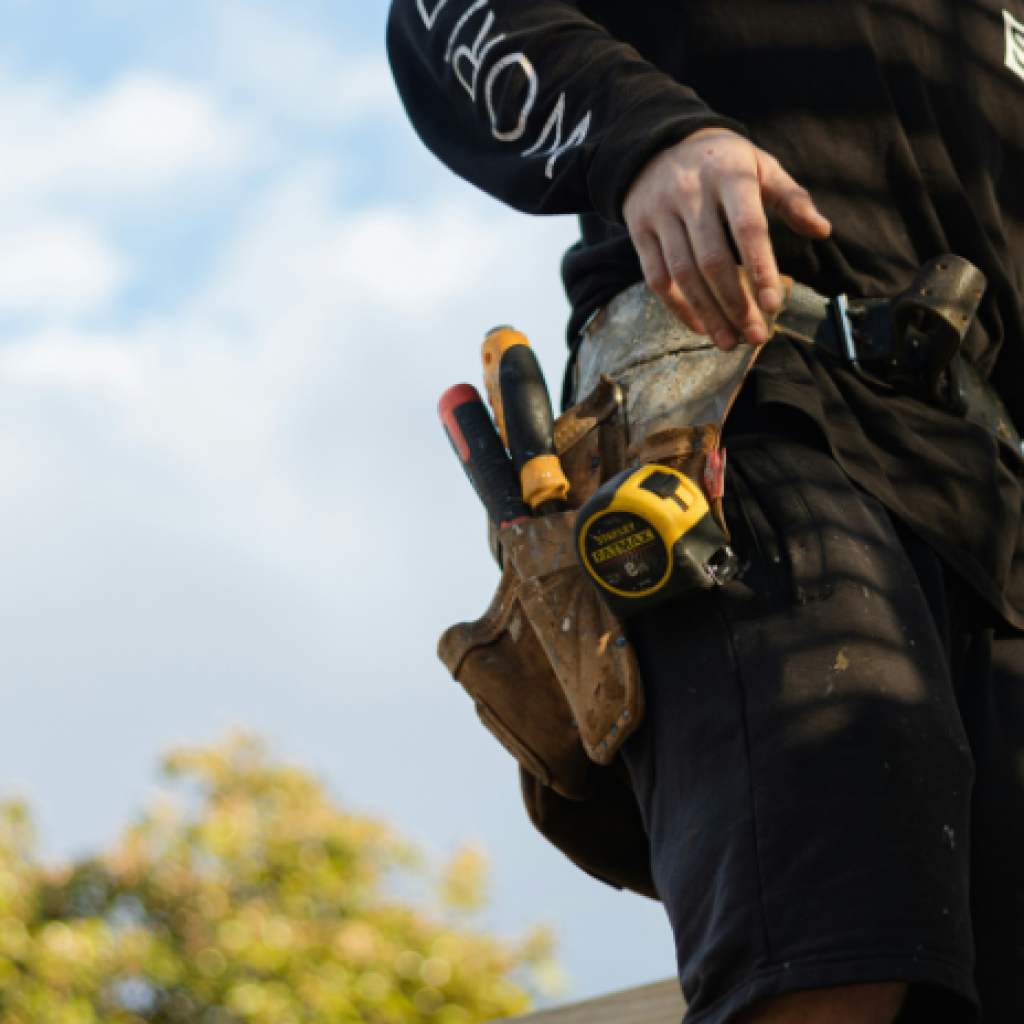 A builder seen from the shoulders down, reaching for something in their tool belt.
