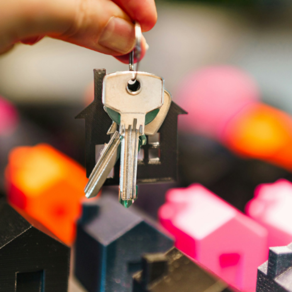 A hand passing house keys over model homes - some pink, some black, some orange, some grey.