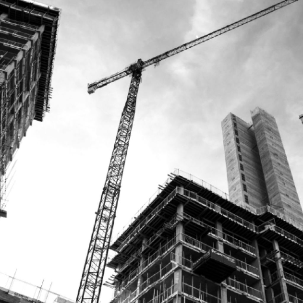 A picture of a city building under construction, looking up from a ground view towards the top of the structure alongside two cranes.