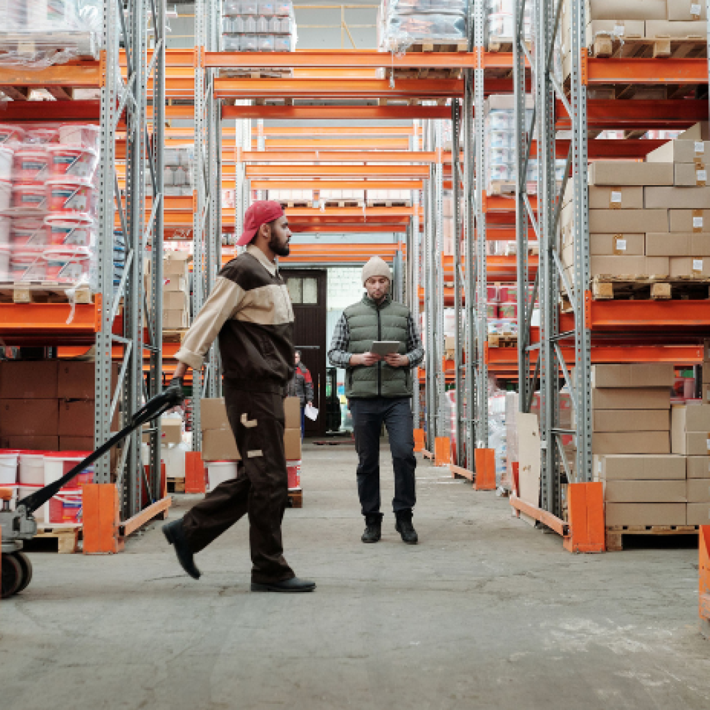 A warehouse employee assessing a temporary warehouse worker hauling inventory on a trolley.