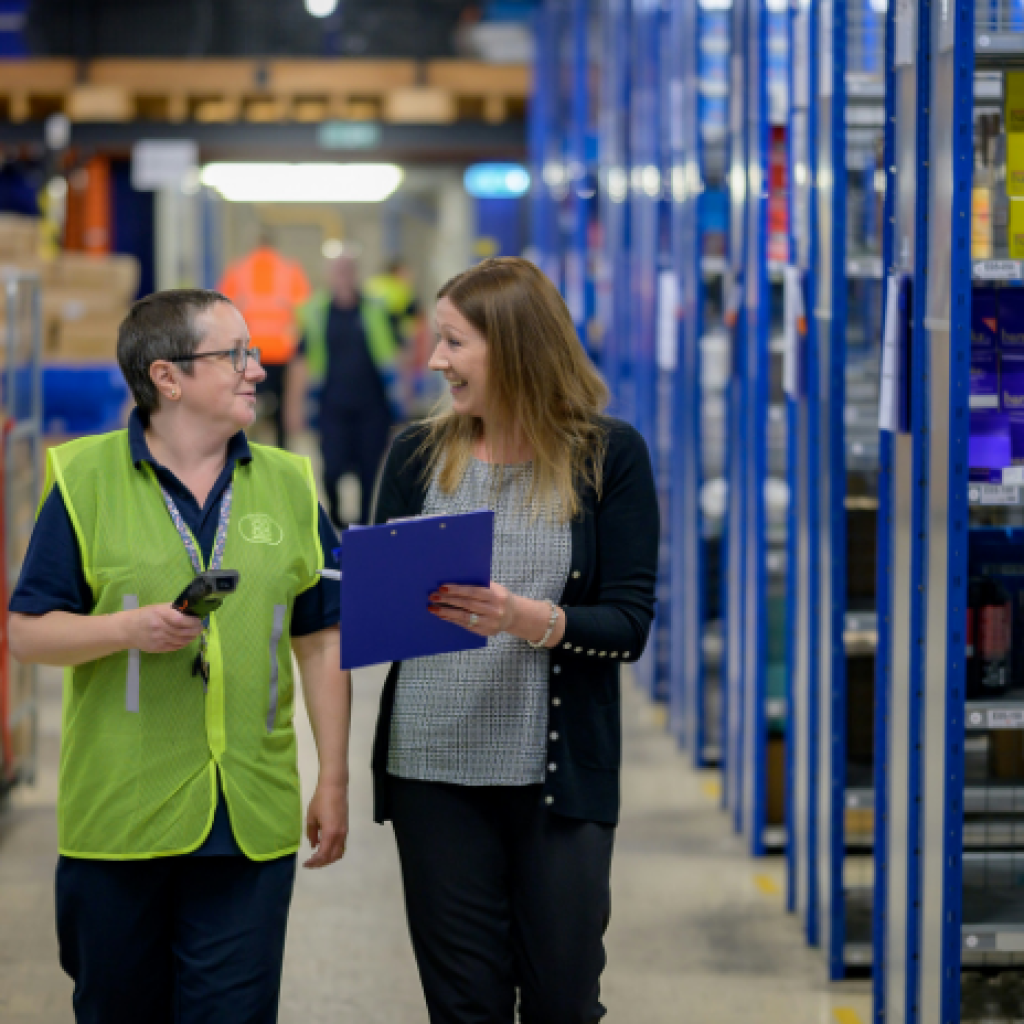 Two people walking through a warehouse - discussing the information on a clipboard that one is carrying.
